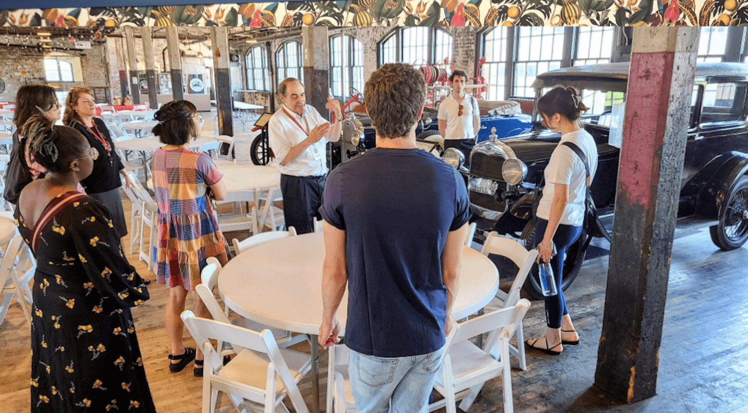 Students standing around a table while a faculty member is speaking. They are in a large warehouse-like room with several cars from the early 20th century. 