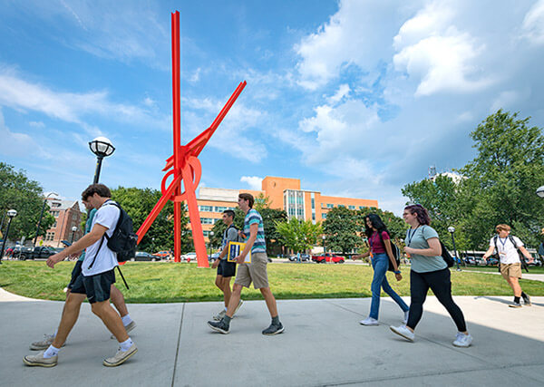 Students walking past the bright red sculpture in front of the UMMA with the LSA Building in the background